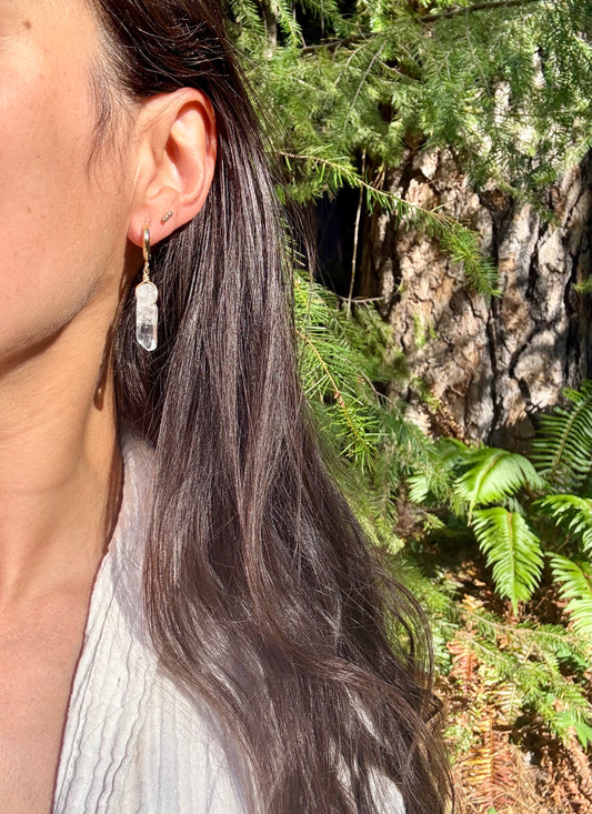 A close-up side profile of a woman with long dark hair wearing a dangling clear crystal earring, set against a natural outdoor background of green ferns and a tree trunk.